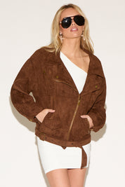 A woman in a white dress and brown jacket poses confidently against a plain background. She wears sunglasses.