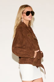 A woman in a white dress and brown jacket poses confidently against a plain background. She wears sunglasses and tan lace-up boots.