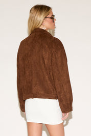 A woman in a white dress and brown jacket poses confidently against a plain background. She wears sunglasses 