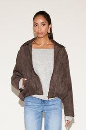 Model wearing a cropped brown suede-look jacket layered over a light gray long-sleeve top, paired with light wash jeans, photographed against a neutral background.
