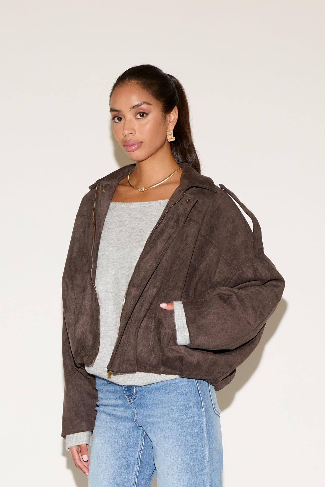 Model wearing a cropped brown suede-look jacket layered over a light gray long-sleeve top, paired with light wash jeans, photographed against a neutral background.