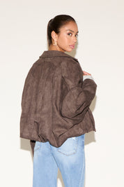 Back view of model wearing a brown suede-look jacket with a relaxed, slightly cropped fit, styled with light wash jeans, photographed against a clean studio background.