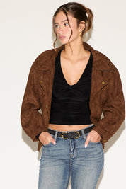 Woman wearing a brown suede jacket, black top, and blue jeans against a white background