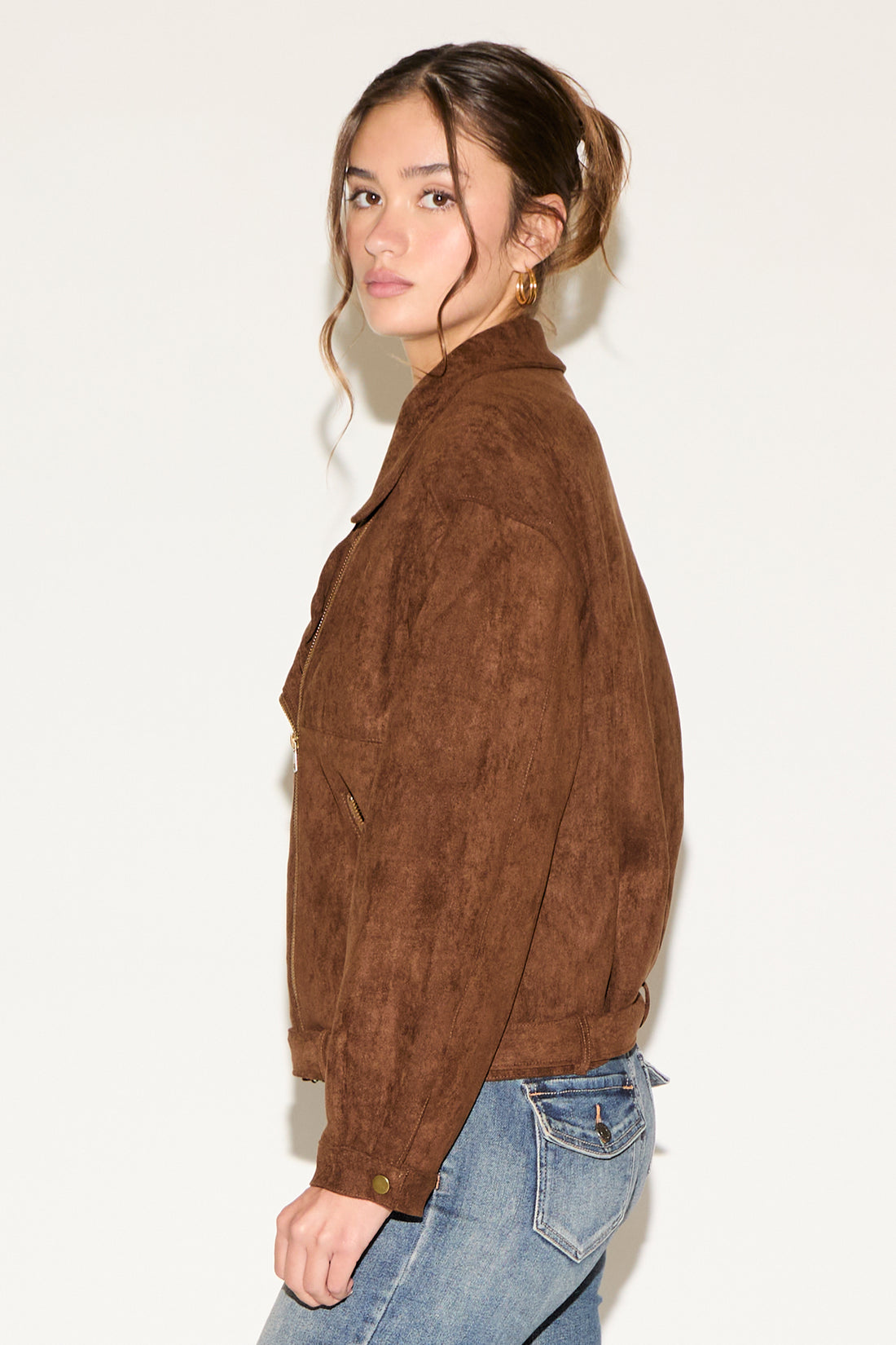 Woman wearing a brown suede jacket, black top, and blue jeans against a white background