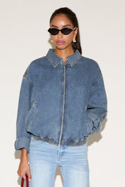 Model wearing a blue denim zip-up jacket with a relaxed fit and elastic hem, styled with light-wash jeans, black oval sunglasses, and gold statement earrings against a neutral background.