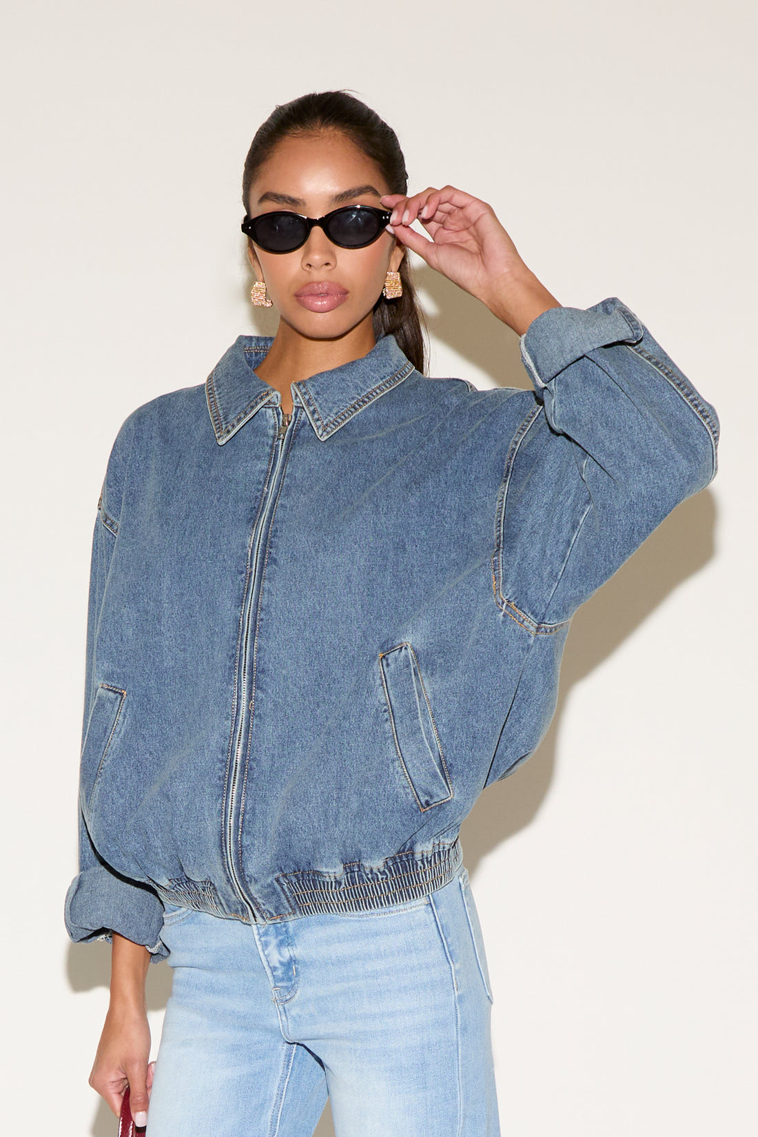 Model wearing a blue denim zip-up jacket with a relaxed fit and elastic hem, styled with light-wash jeans, black oval sunglasses, and gold statement earrings against a neutral background.