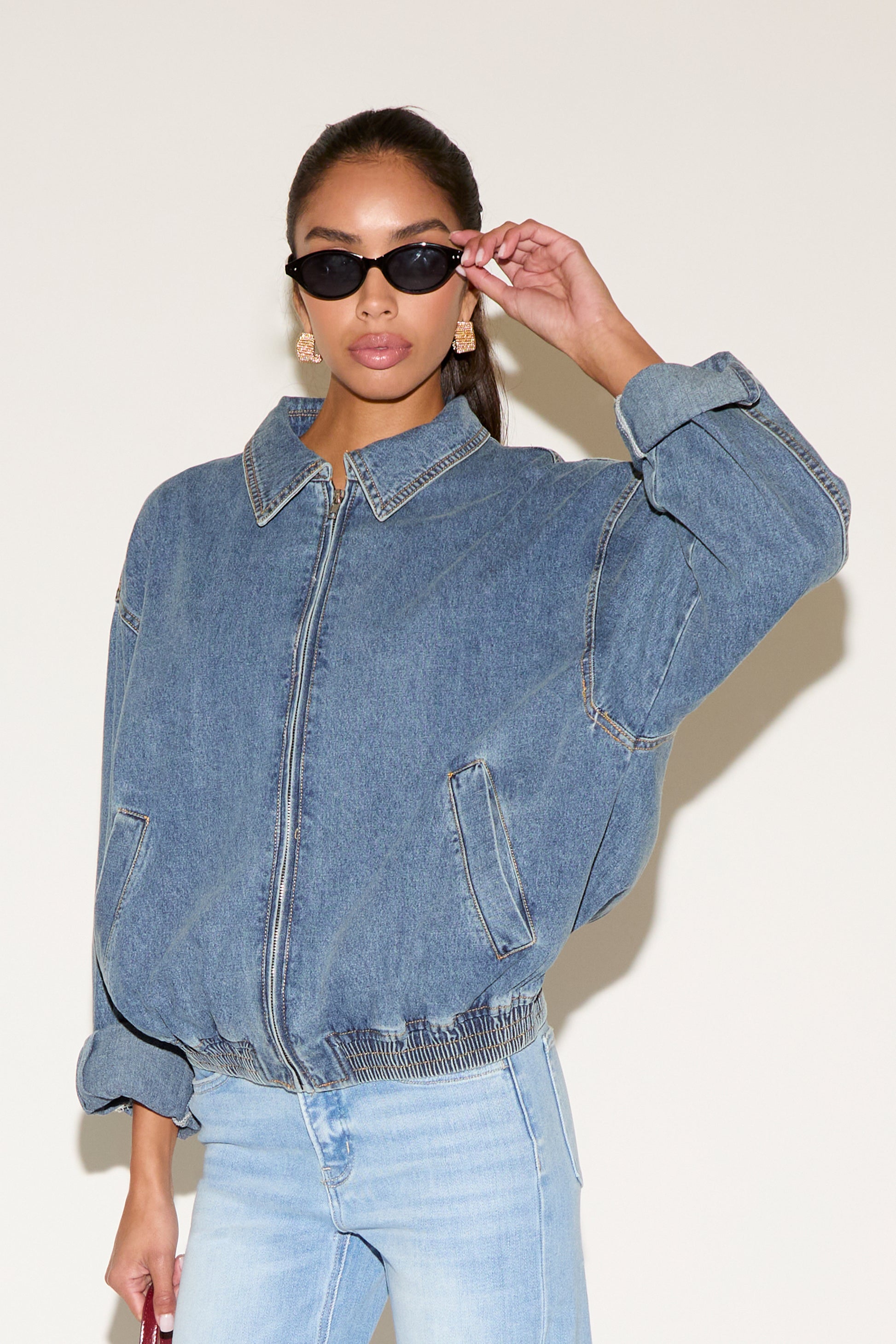 Model wearing a blue denim zip-up jacket with a relaxed fit and elastic hem, styled with light-wash jeans, black oval sunglasses, and gold statement earrings against a neutral background.