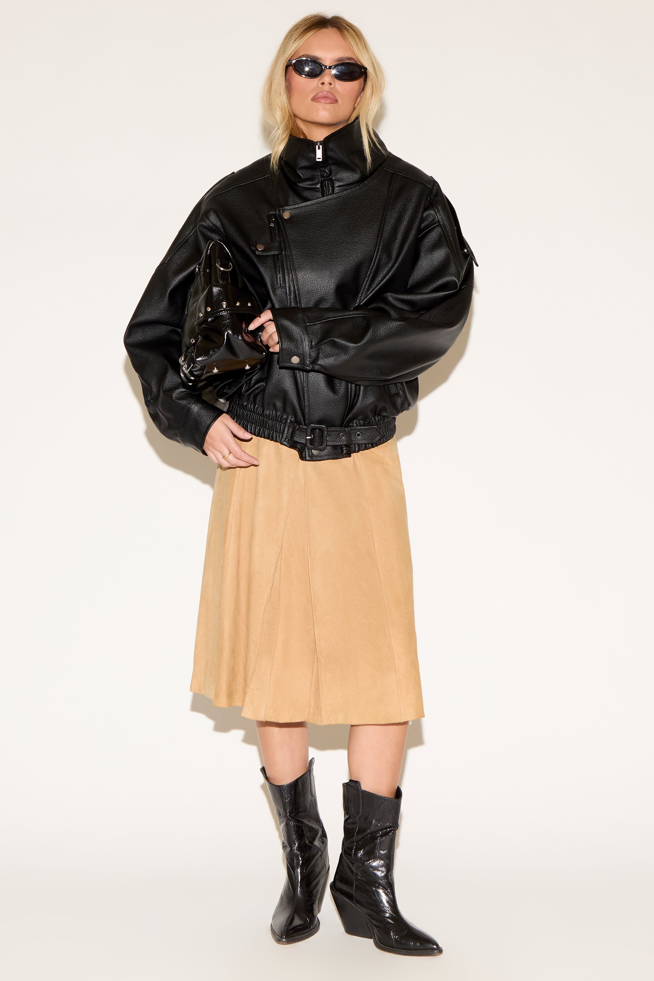 A person wears a black leather jacket, beige skirt, black boots, and sunglasses, exuding a confident, stylish vibe. They hold a shiny black bag.