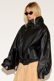 A person wears a black leather jacket, beige skirt, black boots, and sunglasses, exuding a confident, stylish vibe. They hold a shiny black bag.