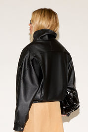 A person wears a black leather jacket, beige skirt, black boots, and sunglasses, exuding a confident, stylish vibe. They hold a shiny black bag.