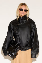 A person wears a black leather jacket, beige skirt, black boots, and sunglasses, exuding a confident, stylish vibe. They hold a shiny black bag.