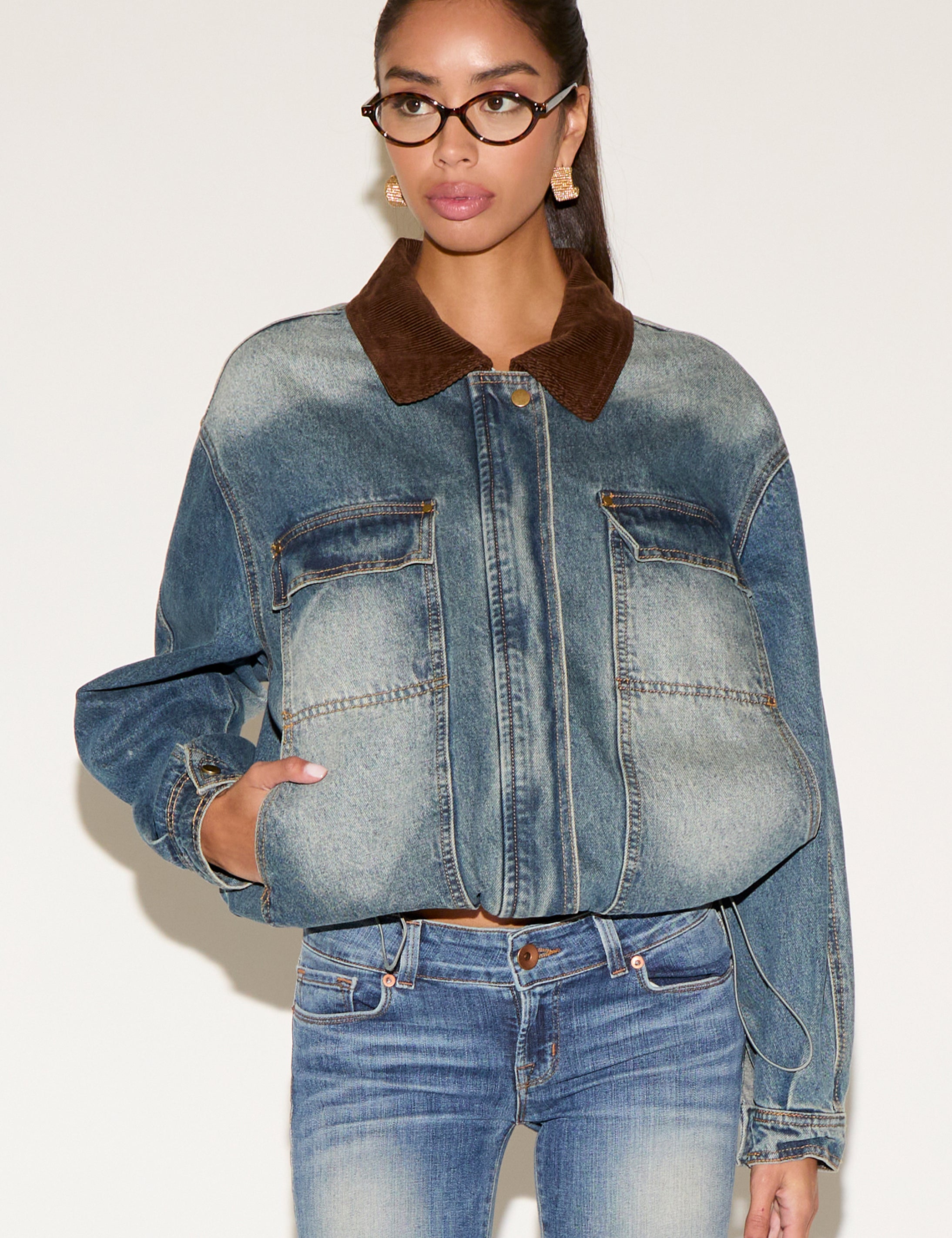 Model wearing a blue denim jacket with a contrast brown corduroy collar, styled with matching denim jeans, tortoiseshell glasses, and gold statement earrings, seated against a neutral background.