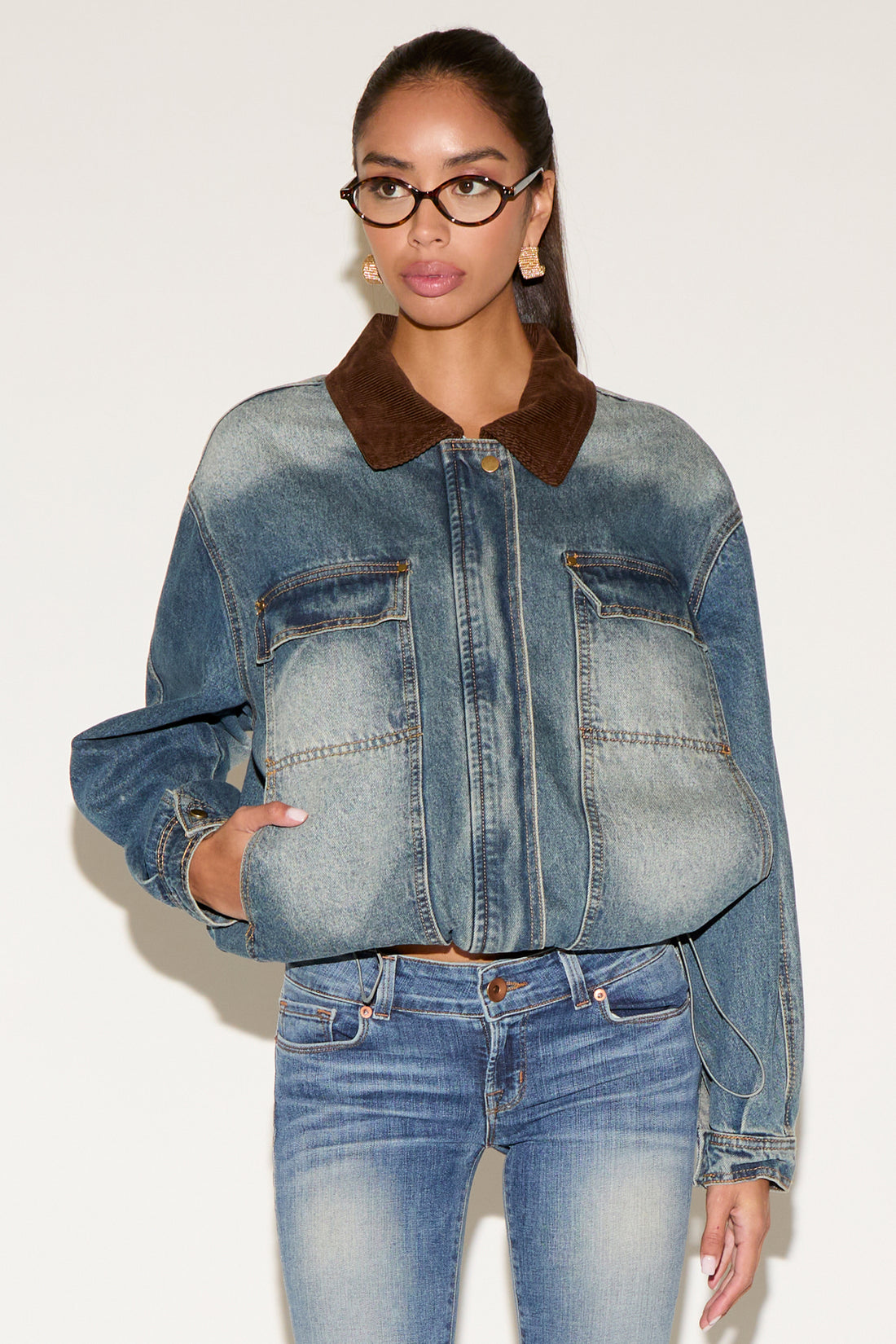 Model wearing a blue denim jacket with a contrast brown corduroy collar, styled with matching denim jeans, tortoiseshell glasses, and gold statement earrings, seated against a neutral background.