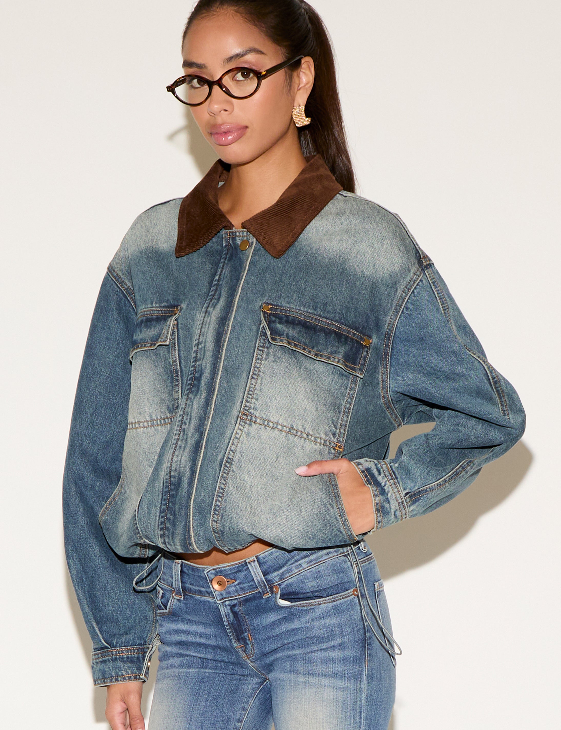 Model wearing a blue denim jacket with a contrast brown corduroy collar, styled with matching denim jeans, tortoiseshell glasses, and gold statement earrings, seated against a neutral background.
