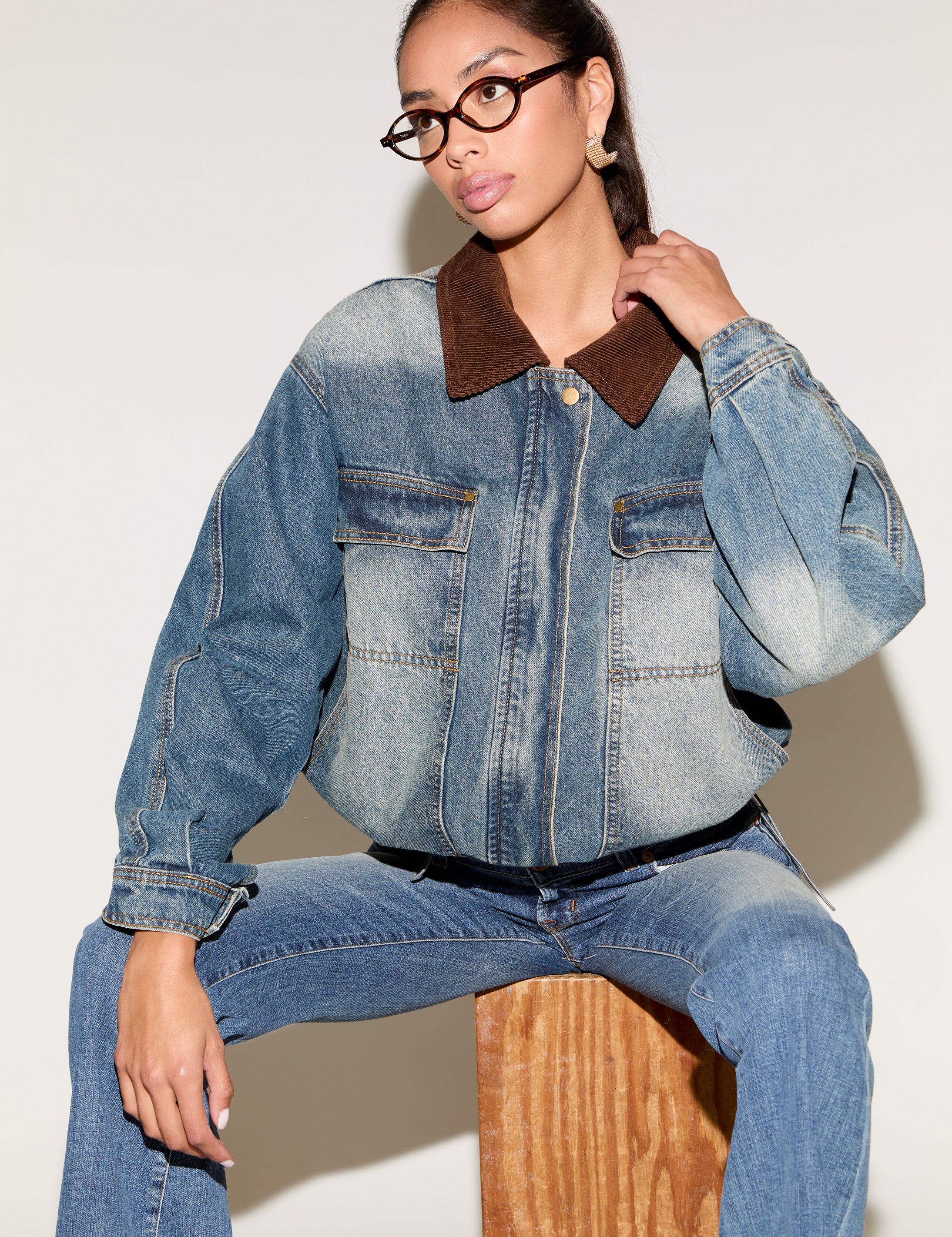 Model wearing a blue denim jacket with a contrast brown corduroy collar, styled with matching denim jeans, tortoiseshell glasses, and gold statement earrings, seated against a neutral background.