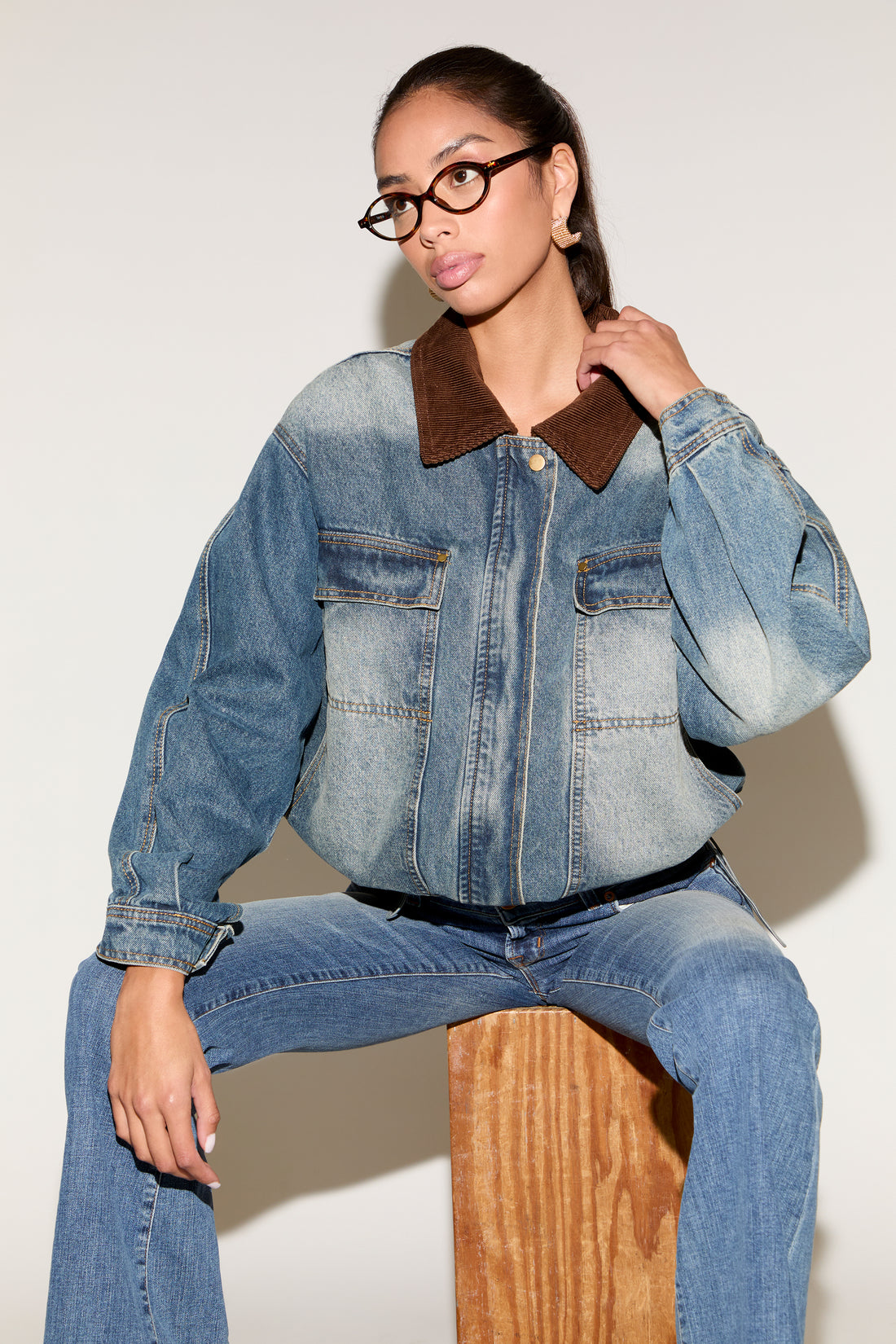 Model wearing a blue denim jacket with a contrast brown corduroy collar, styled with matching denim jeans, tortoiseshell glasses, and gold statement earrings, seated against a neutral background.