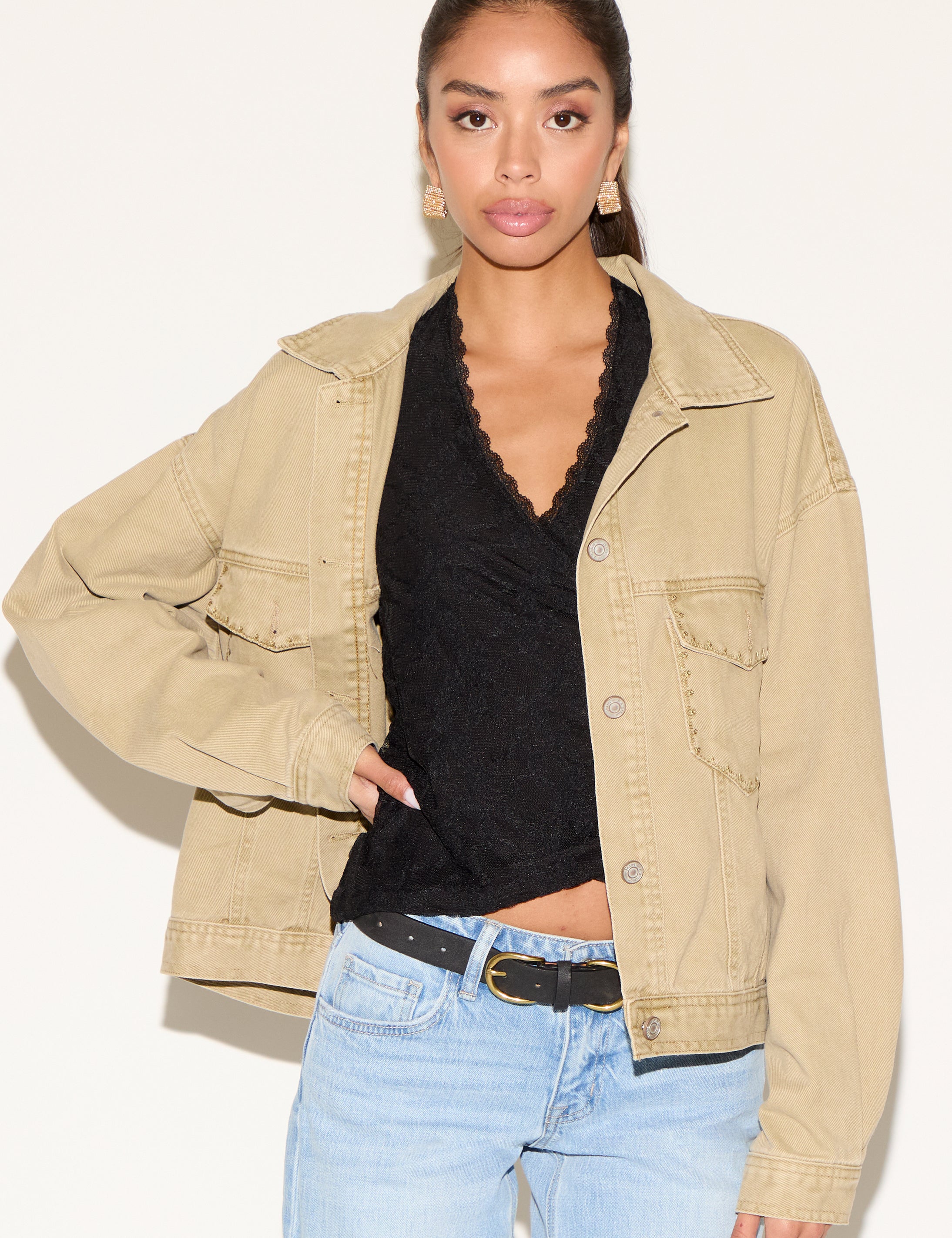 Model wearing a tan corduroy button-up jacket layered over a black lace V-neck top, styled with light-wash denim jeans, a black belt, and gold statement earrings against a neutral background.