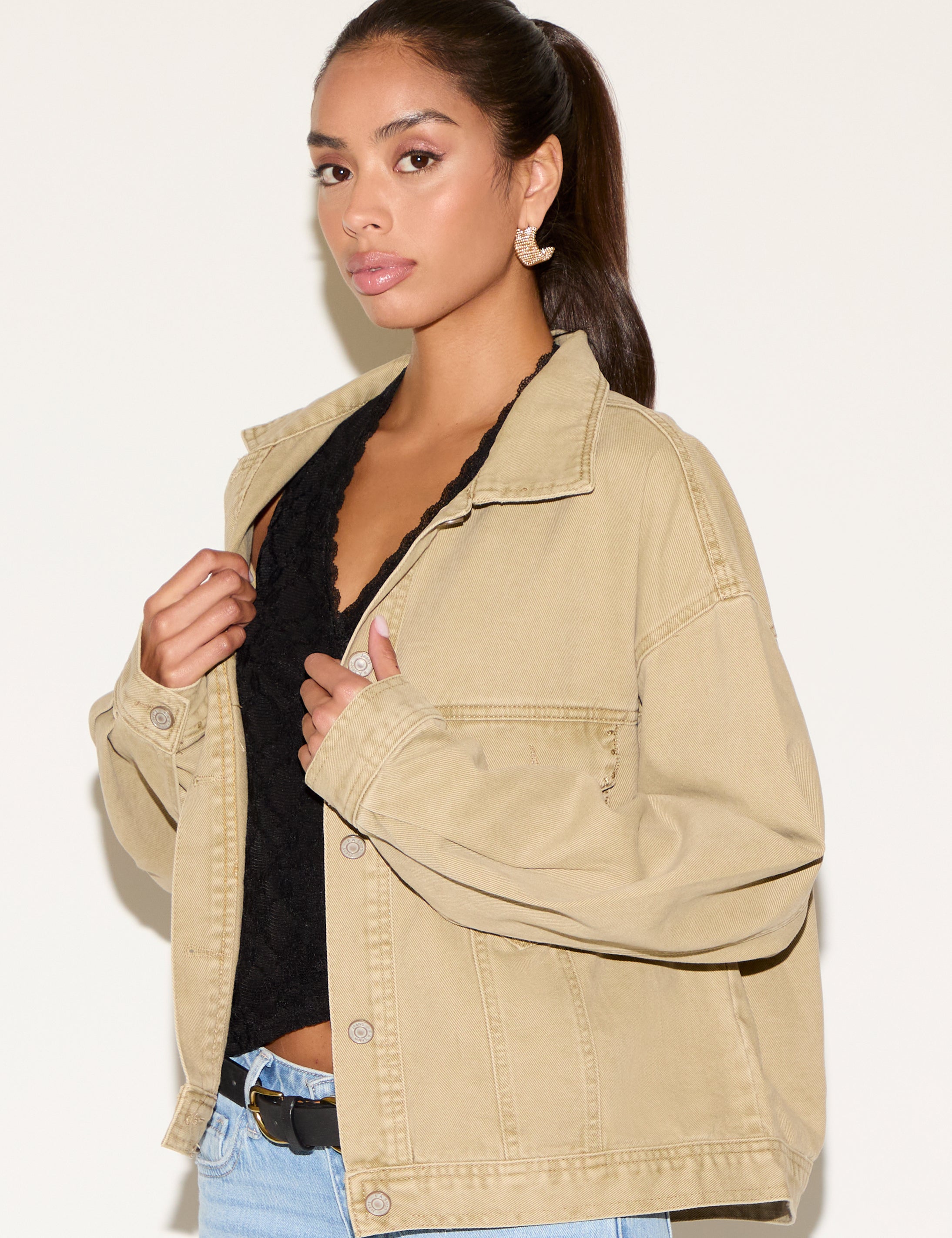 Model wearing a tan corduroy button-up jacket layered over a black lace V-neck top, styled with light-wash denim jeans, a black belt, and gold statement earrings against a neutral background.