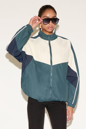 Model wearing a color-block zip-up windbreaker jacket in teal, cream, and navy with a high collar and reflective piping, paired with black leggings and sunglasses, standing against a neutral background.