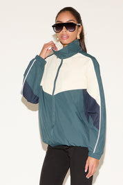 Model wearing a color-block zip-up windbreaker jacket in teal, cream, and navy with a high collar and reflective piping, paired with black leggings and sunglasses, standing against a neutral background.