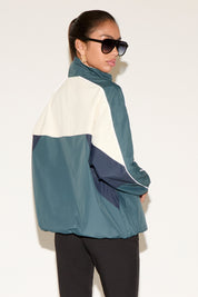Back of model wearing a color-block zip-up windbreaker jacket in teal, cream, and navy with a high collar and reflective piping, paired with black leggings and sunglasses, standing against a neutral background.