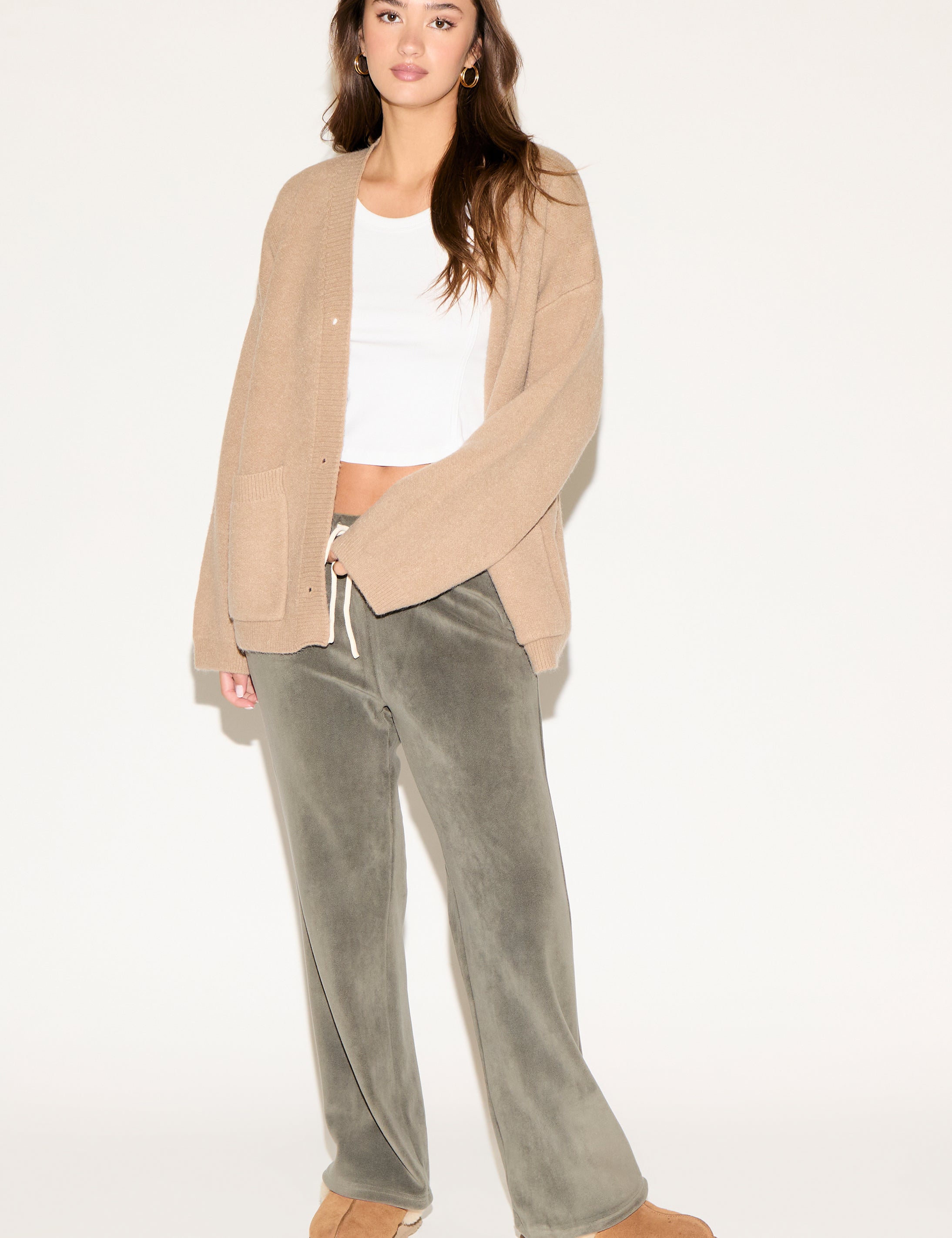 Woman wearing a beige cardigan, white top, and green pants on a white background