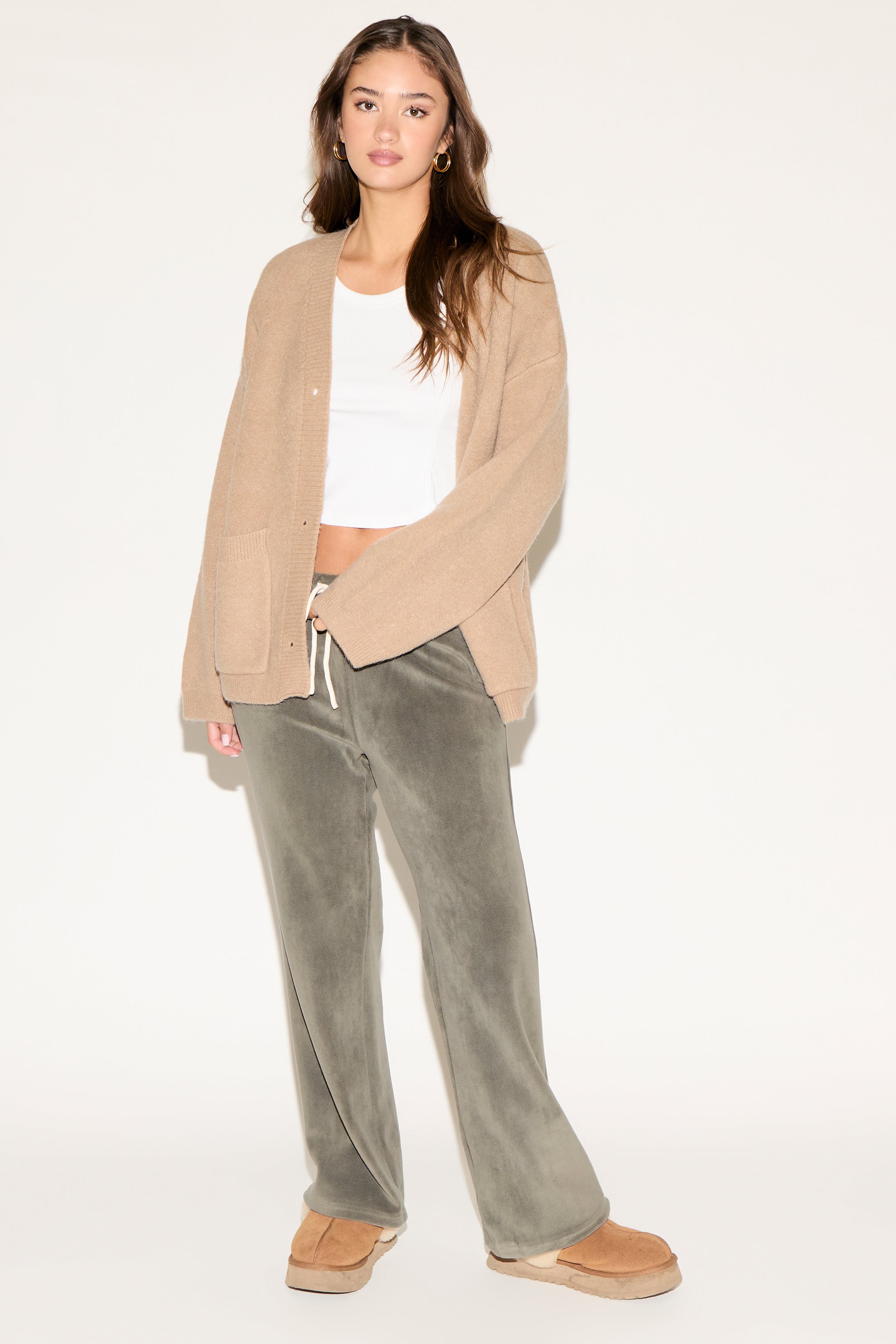 Woman wearing a beige cardigan, white top, and green pants on a white background