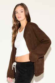 Woman wearing a brown cardigan over a white top and black pants on a plain background