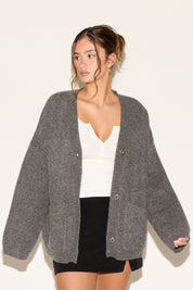 Woman wearing a gray cardigan over a white top and black skirt on a plain background