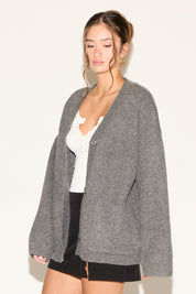 Woman wearing a gray cardigan over a white top and black shorts on a plain background