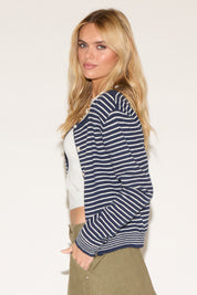 A woman wearing a navy striped cardigan over a white crop top and olive skirt. 