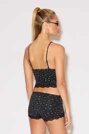 A woman poses confidently in a floral-patterned black lace-trimmed camisole and shorts. She wears blue-tinted glasses and hoop earrings, exuding a stylish and relaxed vibe.