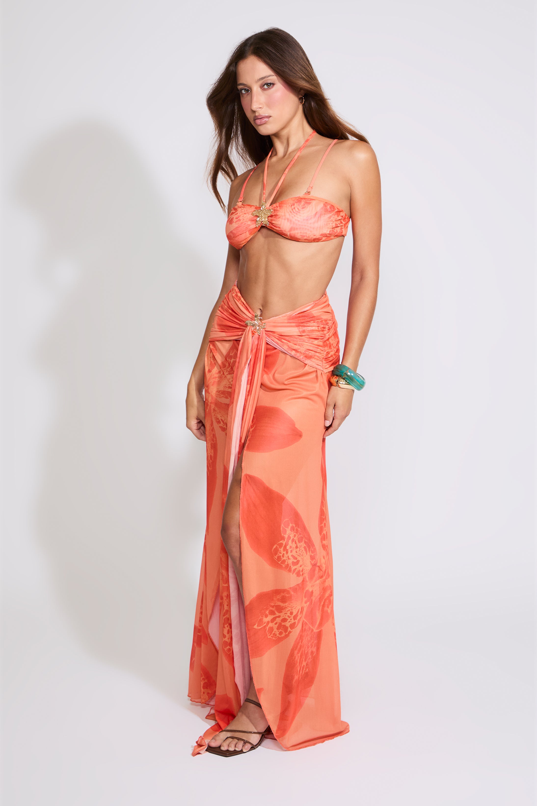 Model is wearing a orange LILY PRINT HALTER BRA TOP & WATERFALL MAXI SKIRT SET with brown heels on a white background.