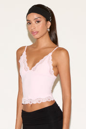 Model wearing a blush pink lace-trim camisole with delicate spaghetti straps and a scalloped hem. Styled with black pants and a black headband and feminine lace details along the neckline, set against a neutral background.
