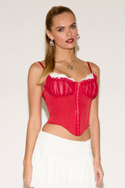 Young woman with long blonde hair, wearing a red corset and a white skirt, poses confidently against a plain background. Her expression is poised and elegant.