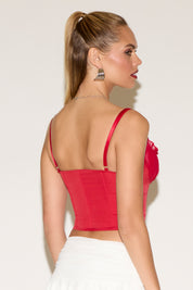 Young woman with long blonde hair, wearing a red corset and a white skirt, poses confidently against a plain background. Her expression is poised and elegant.