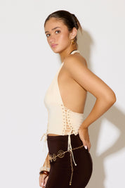 Woman wearing a beige halter top with lace-up details and black pants against a white background