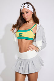 Model is wearing a green a yellow Brazil logo bralette top with a heather grey hoodie and a heather grey pleated skirts on a white background.