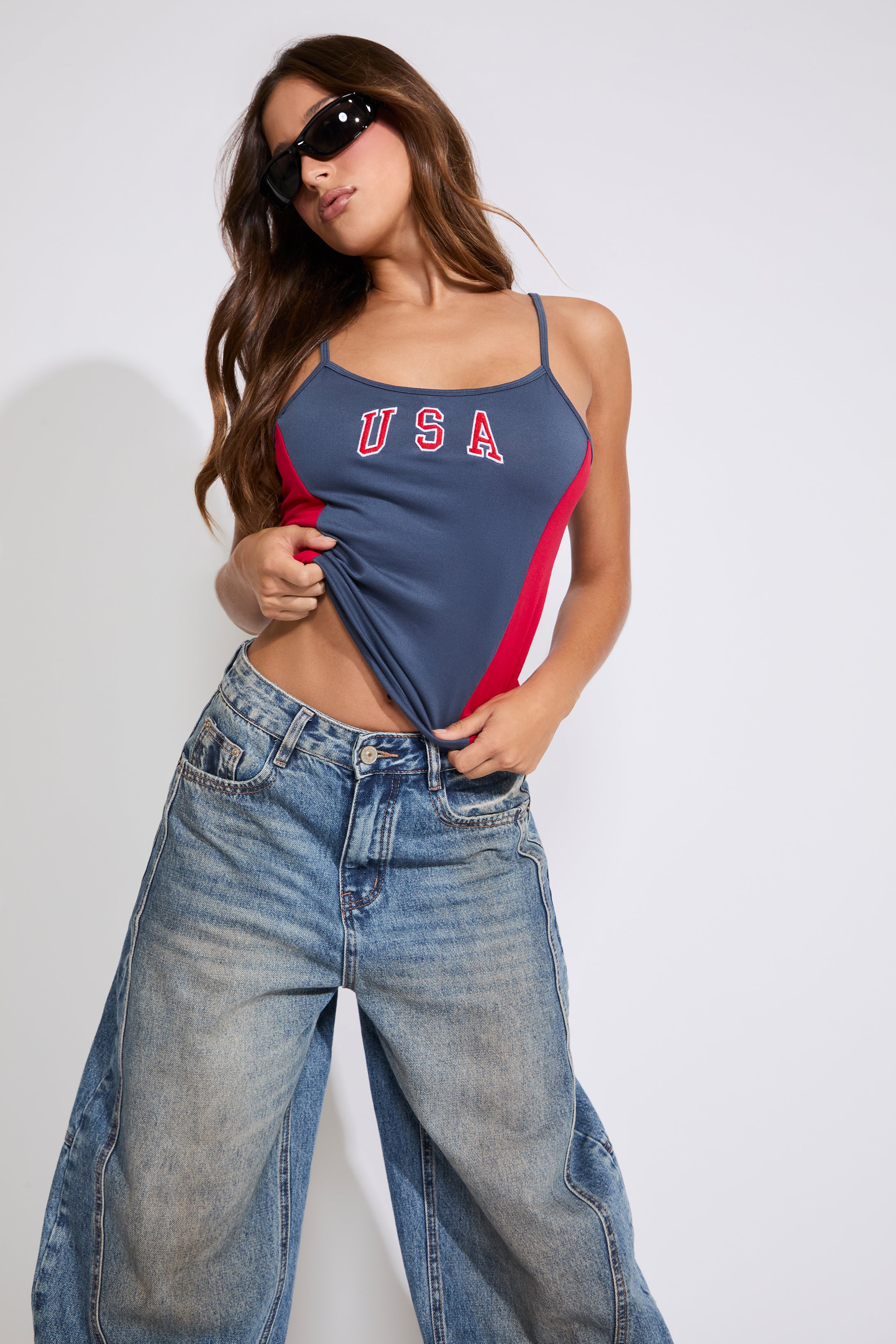 Model is wearing a navy COLOR BLOCK USA & FLAG LOGO SQUARE NECK CAMI TOP with medium denim jeans on a white background. 