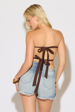 Model is wearing a brown open tie back halter a square neck scarf top with fringe with light denim shorts on a white background.