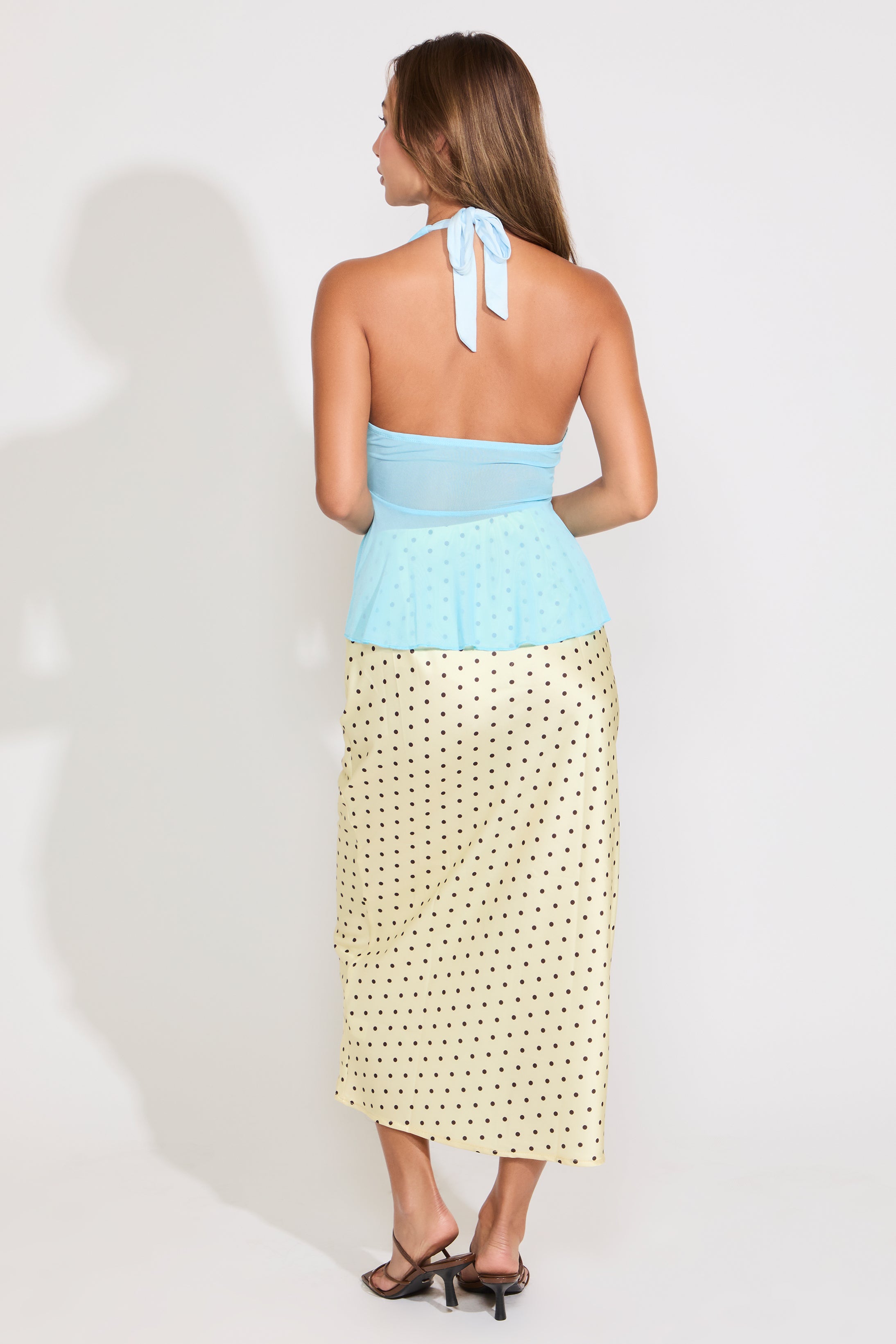 Woman wearing  a sheer blue top with a yellow and black polka dot skirt on a white background.