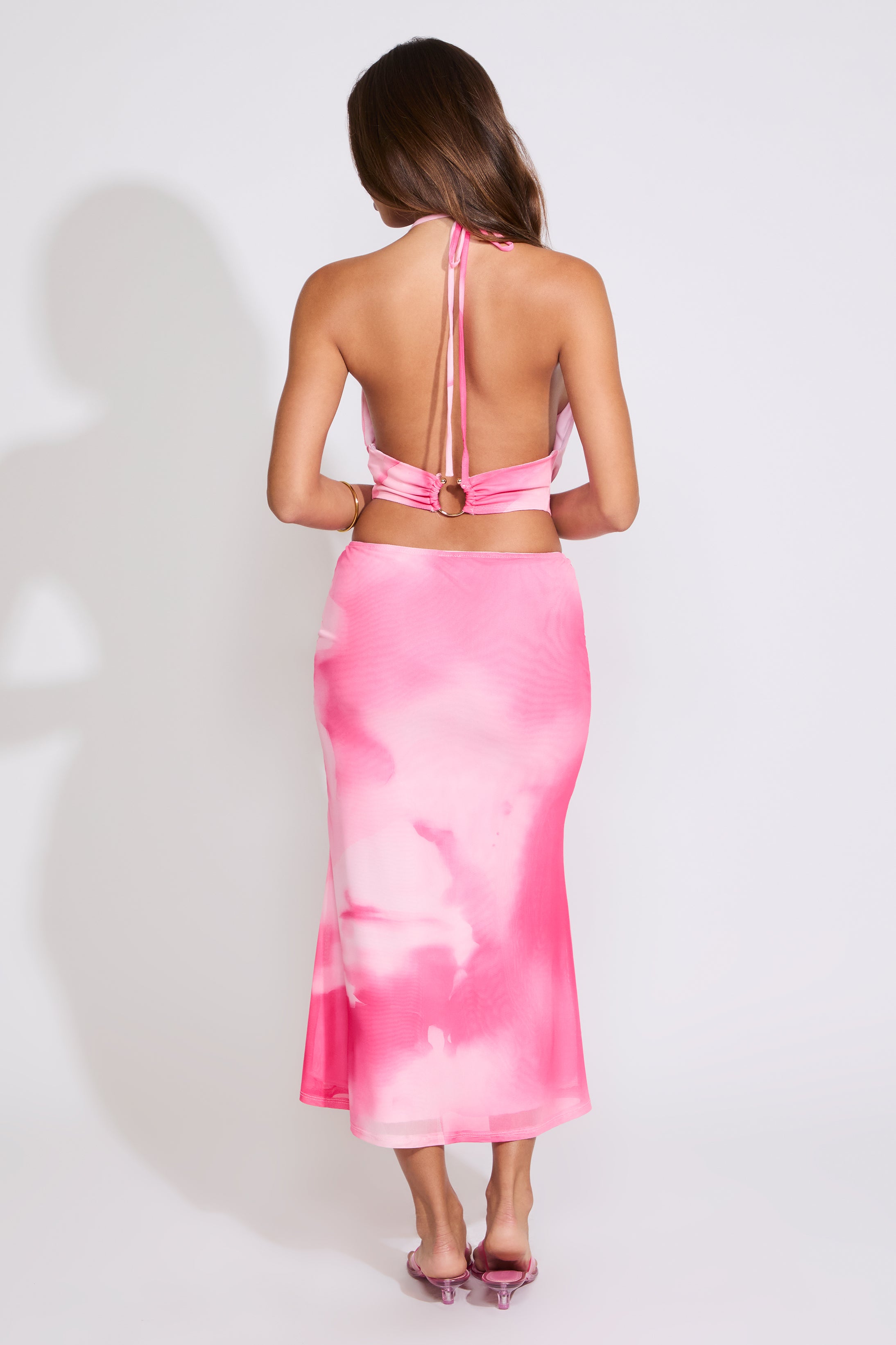 Model is wearing a pink TIE DYE COWL HALTER TOP & MESH MIDI SKIRT SET with pink heels on a white background.