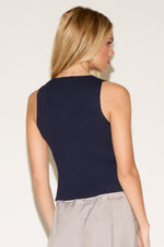 A woman stands confidently against a plain backdrop, wearing a navy sleeveless button-up top and loose, light gray trousers. 