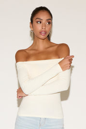 Front view of a model wearing a cream off-the-shoulder long-sleeve top with a smooth, fitted silhouette, styled with light-wash denim. The look highlights a clean neckline and minimal styling, photographed against a neutral studio background.