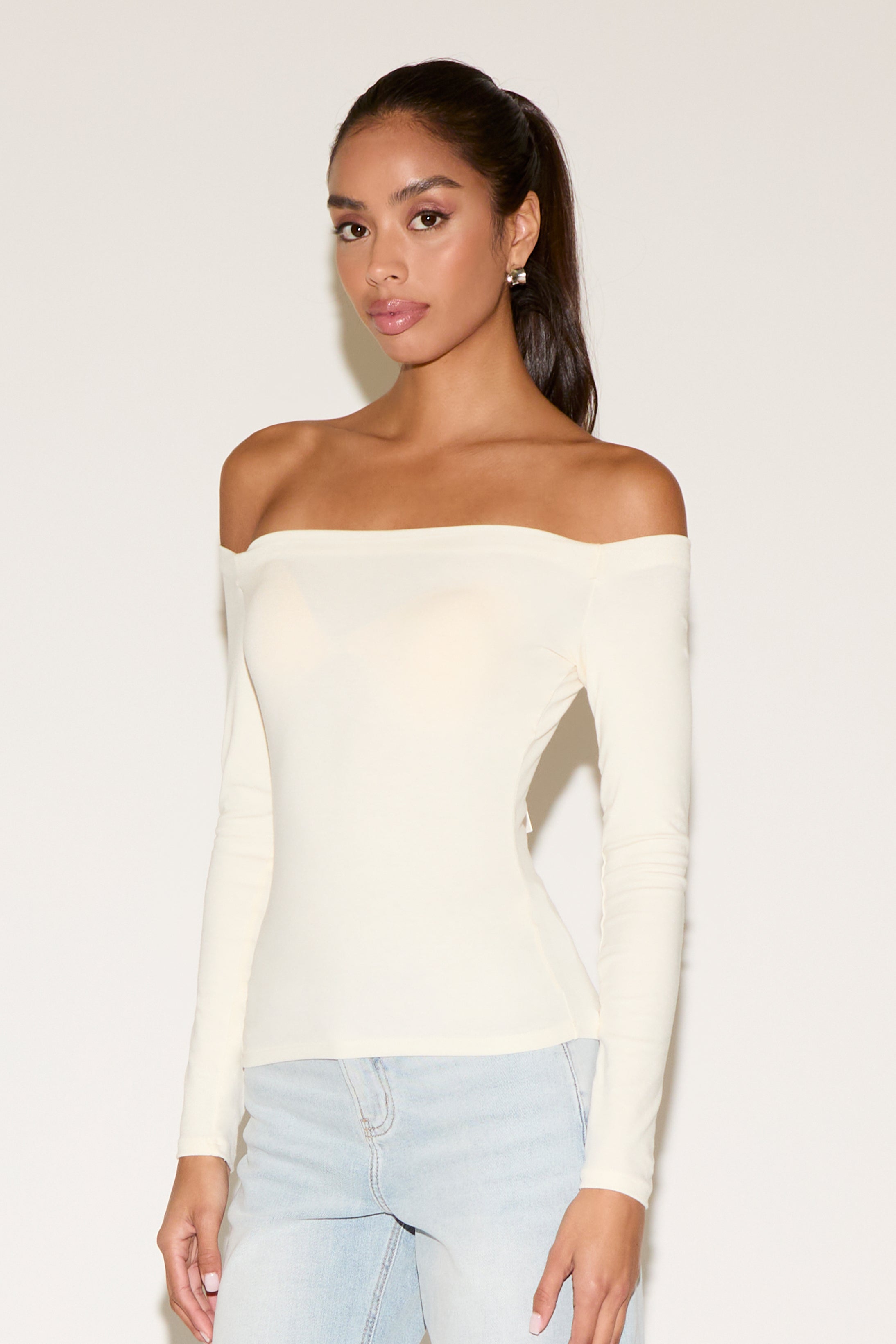 Front view of a model wearing a cream off-the-shoulder long-sleeve fitted top with a sleek, body-hugging silhouette, paired with light-wash denim. The neckline sits straight across the shoulders, creating a clean, minimalist look against a neutral studio background.
