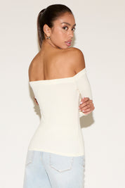 Back view of a model wearing a cream off-the-shoulder long-sleeve fitted top, showcasing a smooth, straight neckline across the shoulders and a flattering, body-contouring fit. Styled with light-wash denim, the look is clean and minimal against a neutral studio background.