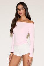 Front view of a model wearing a soft pink off-the-shoulder long-sleeve fitted top with a clean, straight neckline. The top hugs the body for a smooth, flattering silhouette and is styled with white textured shorts. Shot against a neutral studio background for a minimal, polished look.