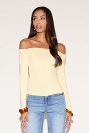 Front-facing studio image of a model wearing a soft yellow off-the-shoulder long-sleeve top with a slim, body-hugging fit. Paired with light-wash high-rise jeans and amber resin bangles, the look is clean and minimal against a neutral background, highlighting the neckline and smooth silhouette.