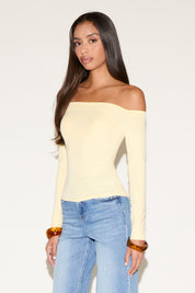 Front view of a model wearing a pale yellow off-the-shoulder long-sleeve fitted top with a smooth, body-contouring silhouette. Styled with light-wash straight-leg jeans and amber bangles, photographed against a clean studio backdrop for a soft, modern look.