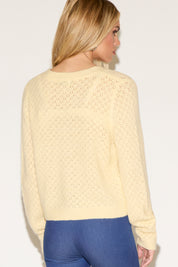 Woman with long blonde hair wearing a yellow buttoned cardigan and blue jeans standing confidently with hands on hips against a plain background.